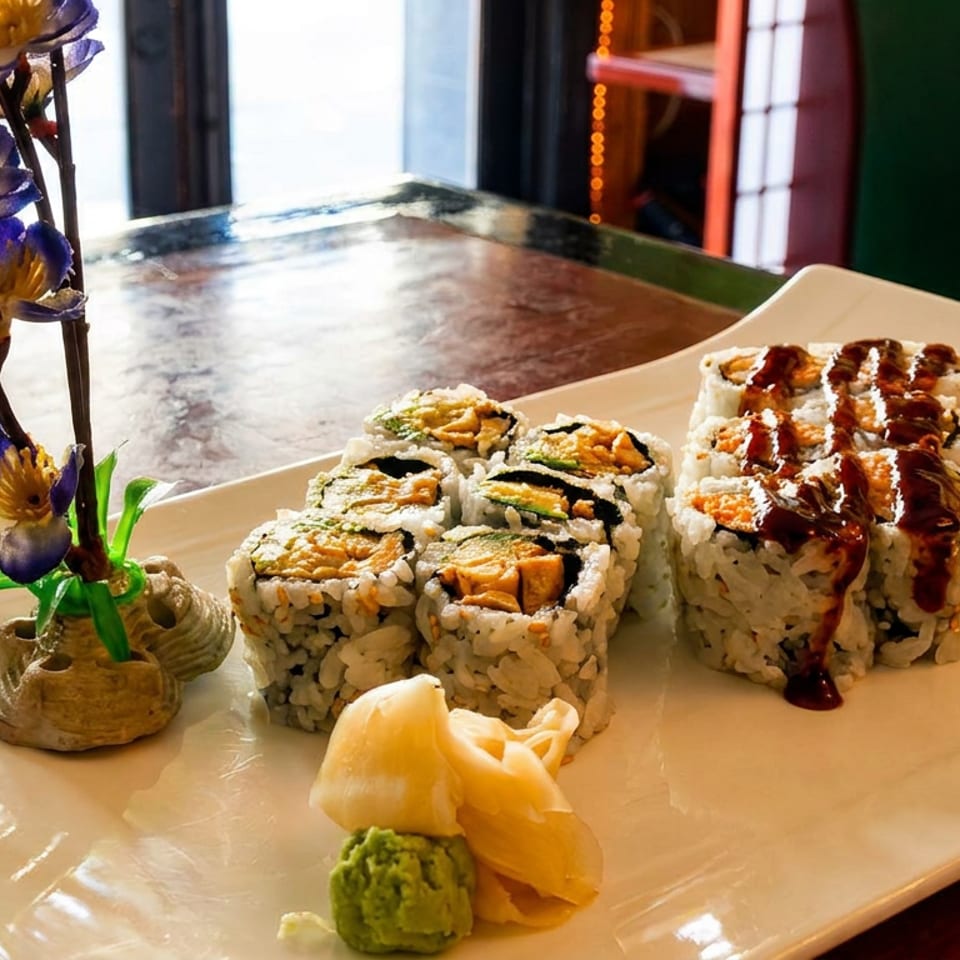 Two rows of sushi rolls on a white plate with pickled ginger, wasabi, and a floral decoration.