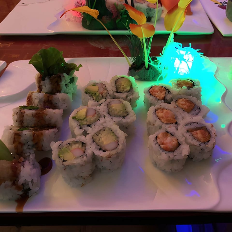 Three rows of sushi rolls with various fillings and sauces on a white plate with colorful lighting.
